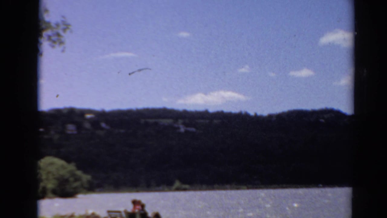 1964 Toledo, Ohio 8mm frame showing a lake, dark wooded hills, scattered clouds, and distant people seated near the shore.