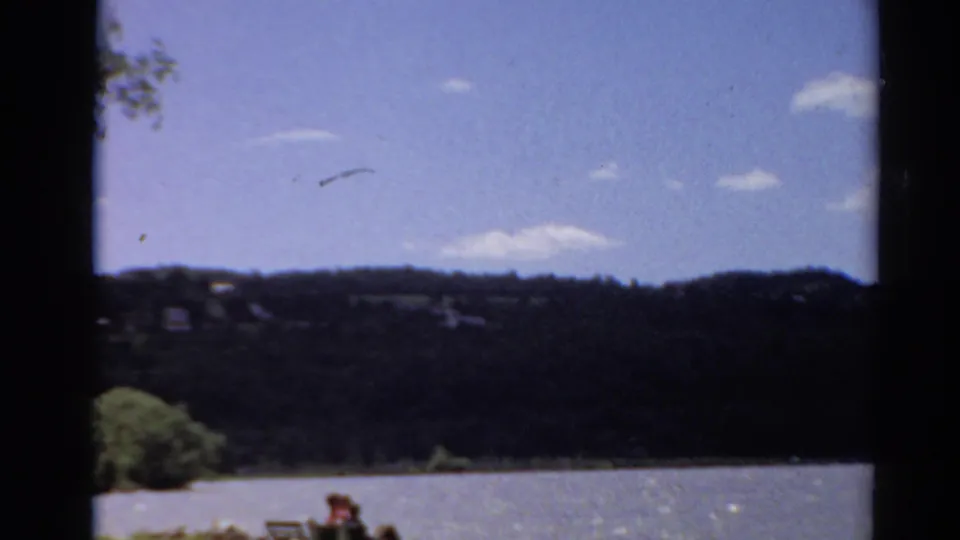 1964 Toledo, Ohio 8mm frame showing a lake, dark wooded hills, scattered clouds, and distant people seated near the shore.
