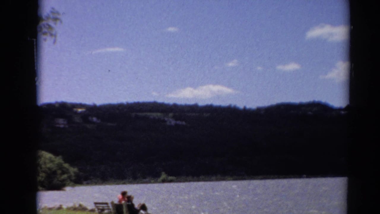 1964 Toledo, Ohio home-movie frame with lake water, wooded ridge, blue sky, and small figures by a shoreline bench.