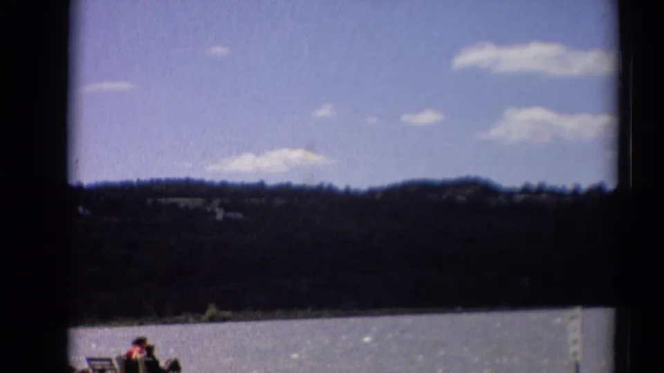 1964 Toledo, Ohio frame showing a soft-focus lake scene with wooded hills, scattered clouds, and distant people at the shore.