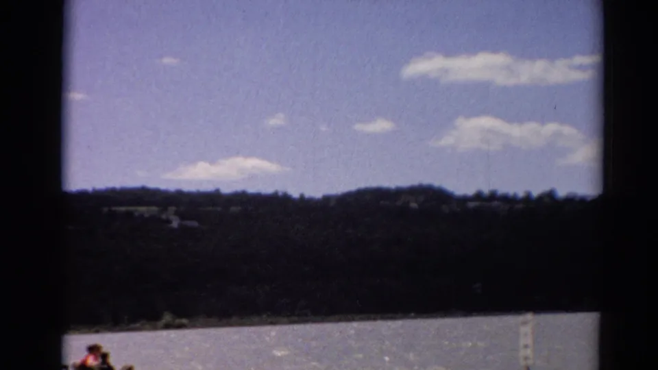 1964 Toledo, Ohio 8mm still with lake water, wooded hills, blue sky, clouds, and partially visible people at the bottom edge.