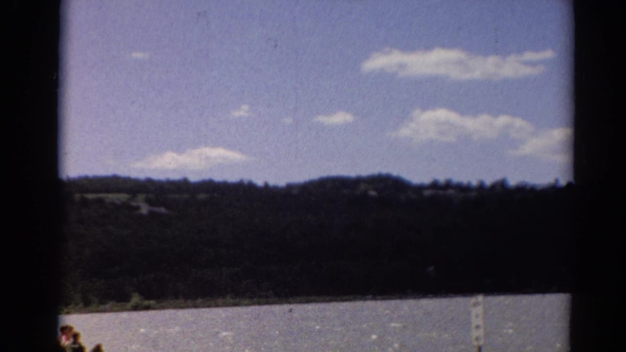 Lake water and a dark tree-lined shore beneath a blue sky, framed by black film borders.