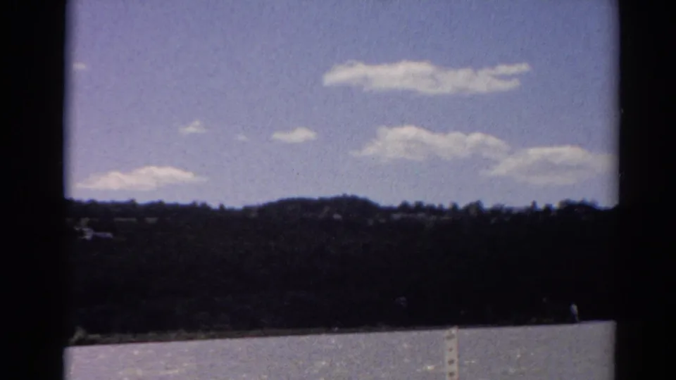 1964 Toledo-area 8mm view across calm water toward a wooded horizon, with scattered clouds and heavy frame edges.