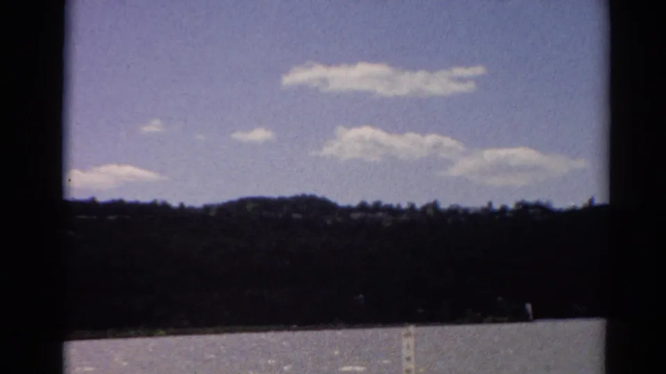 1964 Toledo-area home-movie frame looking over lake water to a dark tree line and clouded summer sky.