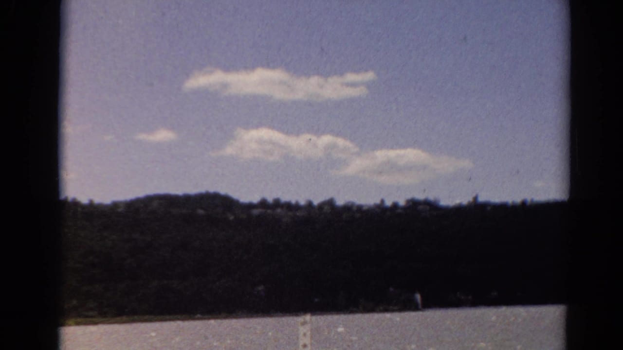 1964 Toledo home-movie frame showing a distant tree-lined shore beneath scattered clouds.