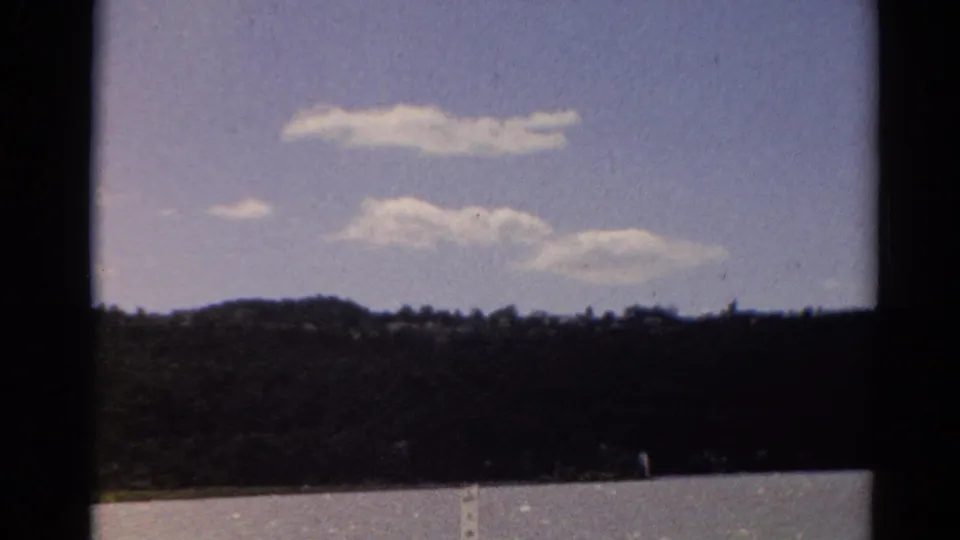 1964 Toledo home-movie frame showing a distant tree-lined shore beneath scattered clouds.