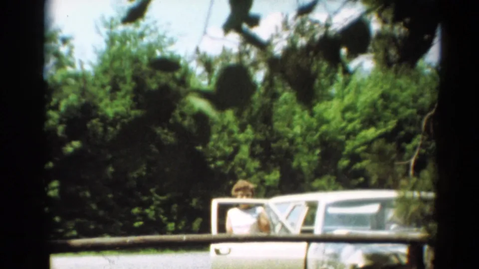 A woman in a light top appears beside the passenger window of a white car in 1964 Toledo home-movie footage.