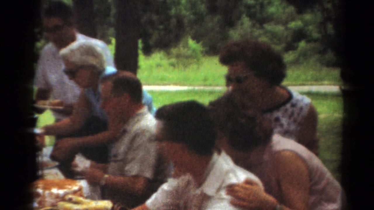 1964 Toledo home-movie frame of adults gathered at a park picnic table, with food spread out and trees and grass behind them.