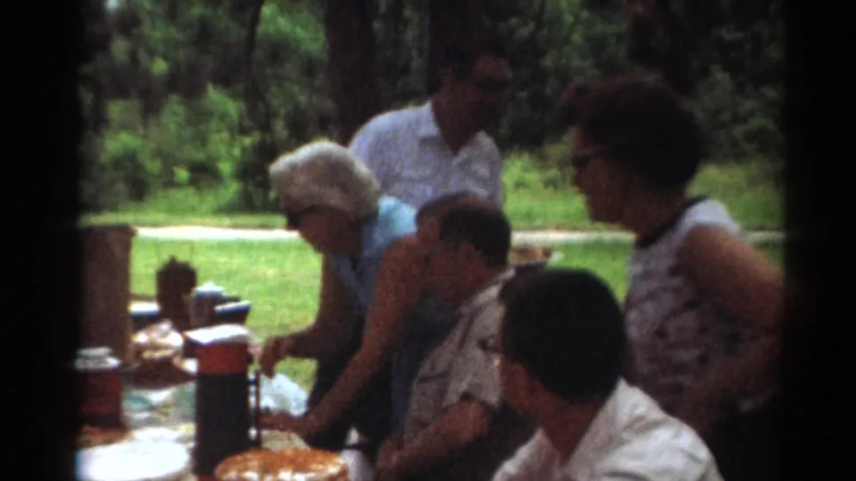 1964 Toledo picnic frame showing adults reaching and standing around a table with food and drink containers in a green park setting.