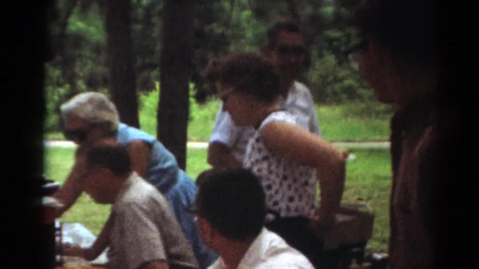 Toledo 1964 home-movie frame of a woman standing by seated adults at a picnic table, with trees and lawn behind the group.