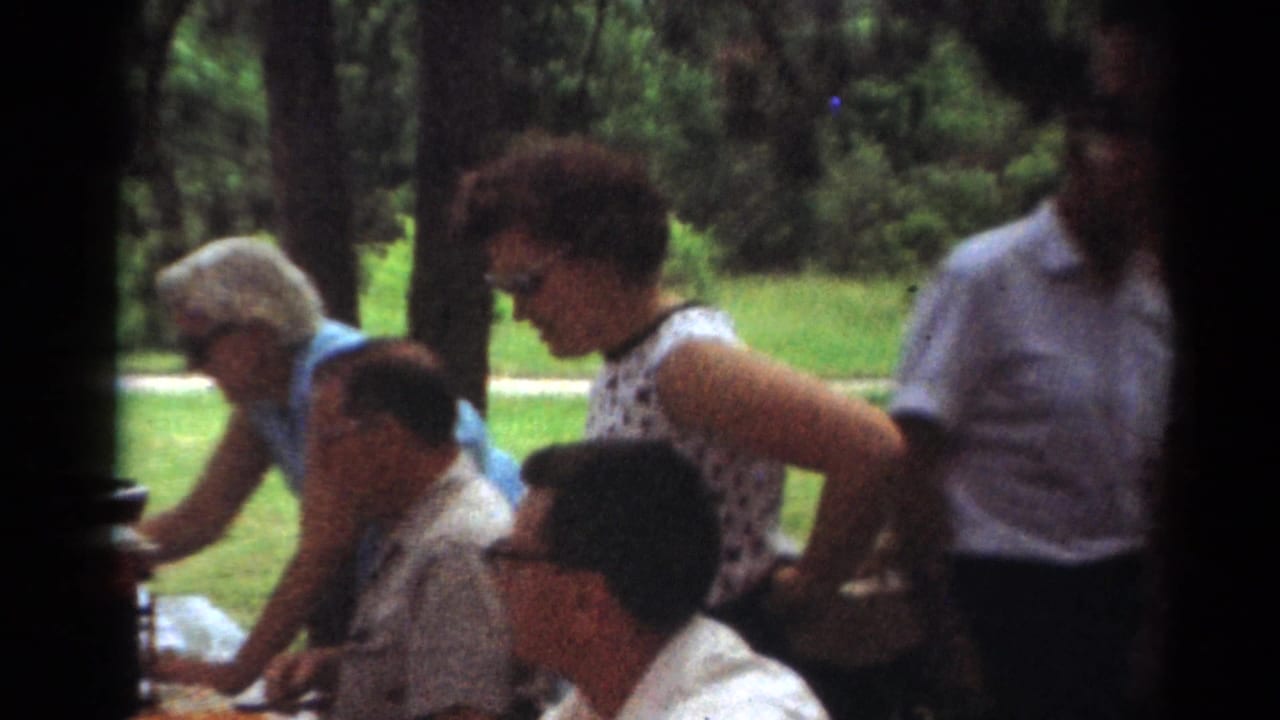 1964 Toledo home-movie frame showing adults in midcentury clothing around a picnic table under trees in a park.