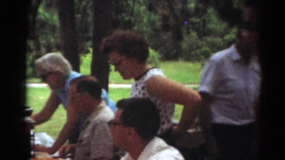 1964 Toledo home-movie frame showing adults in midcentury clothing around a picnic table under trees in a park.