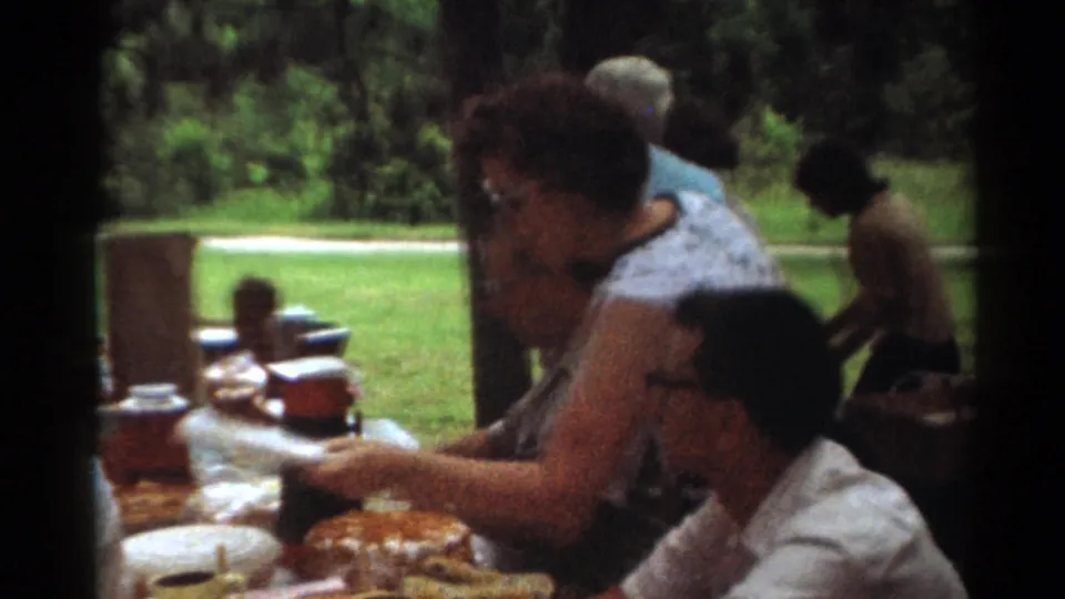 1964 Toledo park picnic frame with adults seated at a table, food containers, thermoses, and grass beyond.