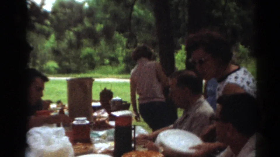 1964 Toledo home-movie frame showing a close picnic-table view with adults, serving dishes, and wooded park background.