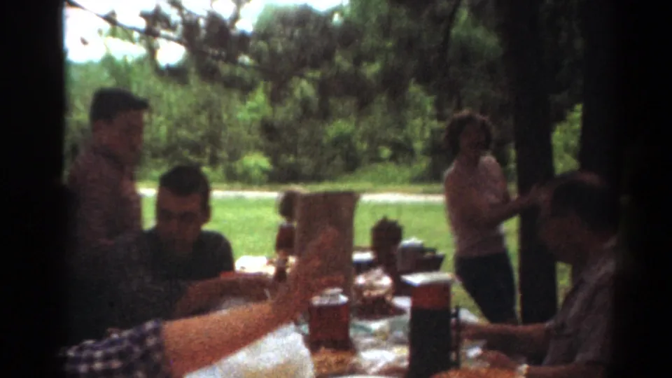 1964 Toledo park picnic frame with adults seated at a table, a standing figure, food items, and shaded greenery.