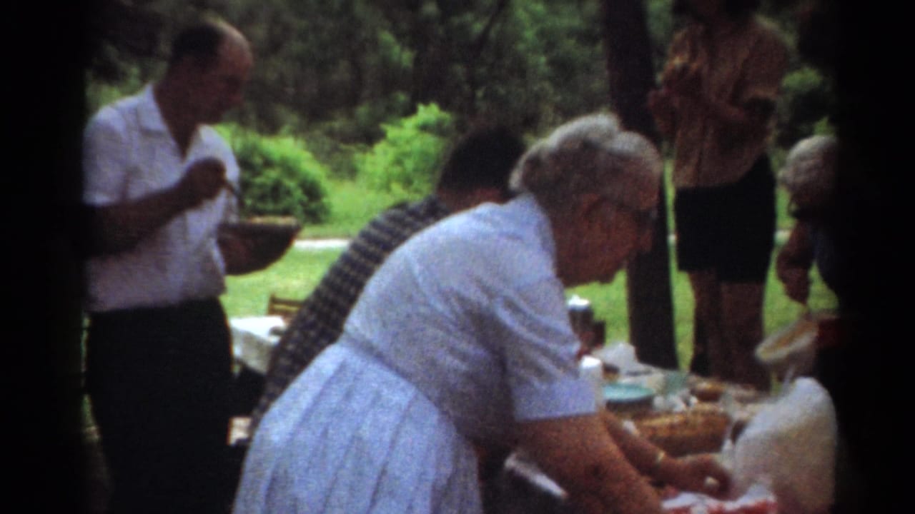 1964 Toledo home-movie frame showing older adults near a picnic table, with a woman in a light dress serving food under trees.