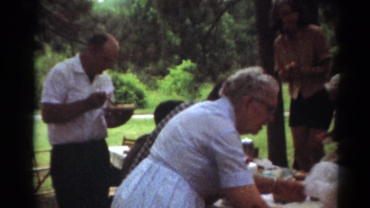 A woman leans over a picnic table while adults stand and eat nearby.