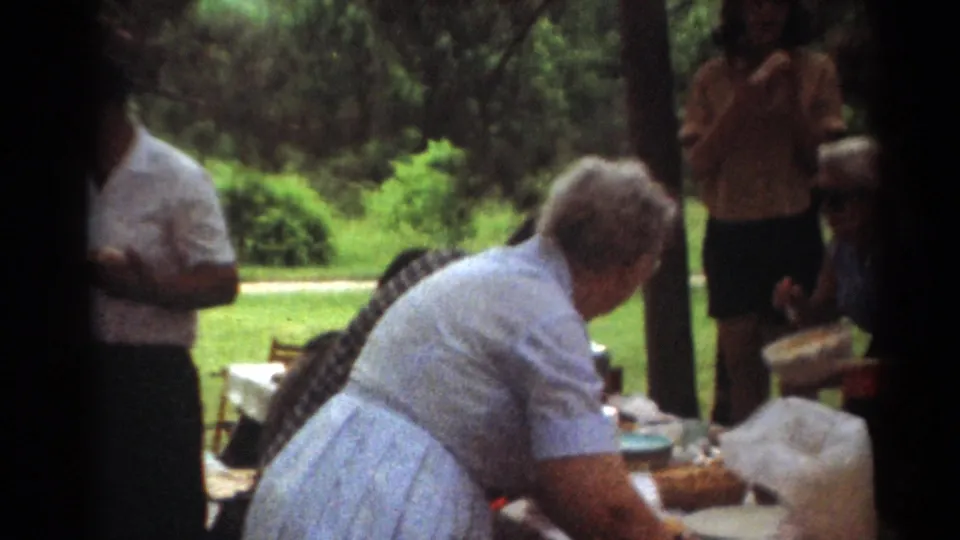 Soft 8mm frame from Toledo in 1964 showing adults at a park picnic table, with food containers and wooded greenery behind them.