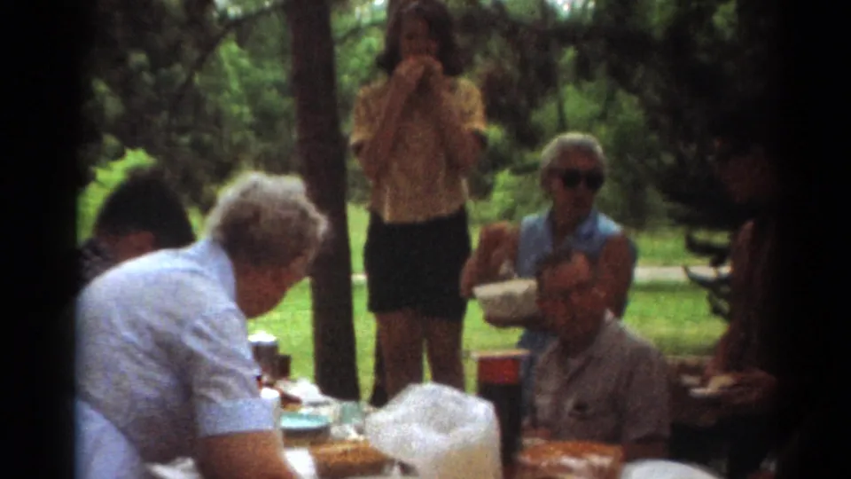 1964 Toledo picnic frame with an older woman at the table, seated adults, food containers, trees, and grass in the background.