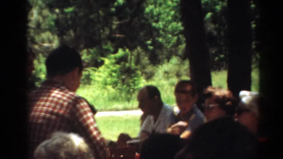 1964 Toledo home-movie frame of adults gathered at a shaded park picnic table.