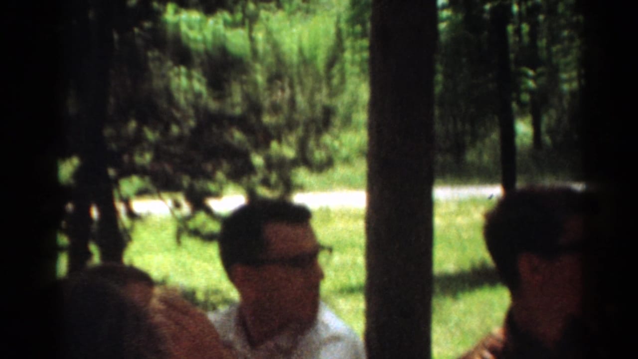 1964 Toledo home-movie frame showing adults at a park picnic table with trees, grass, sunglasses, and dark foreground framing.