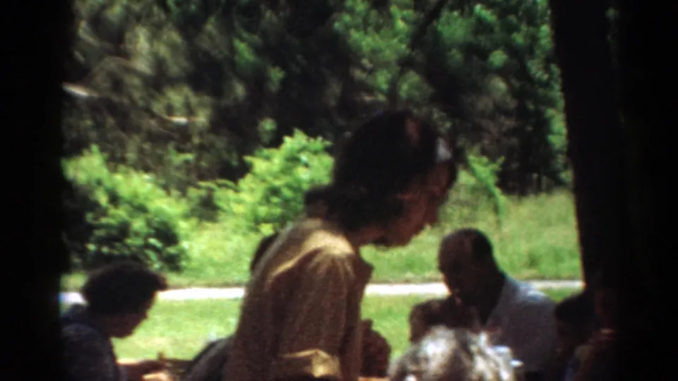 1964 Toledo home-movie still showing a woman leaning near a picnic table while adults sit amid grass, trees, and bright summer light.