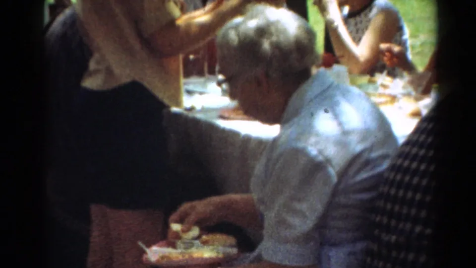 1964 Toledo park picnic frame with an older man eating from a plate, nearby adults, tableware, food, grass, and trees.
