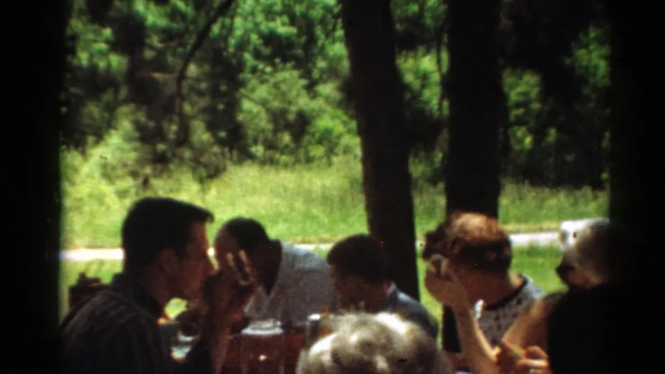 1964 Toledo home-movie frame showing adults eating and talking at a picnic table beneath trees.