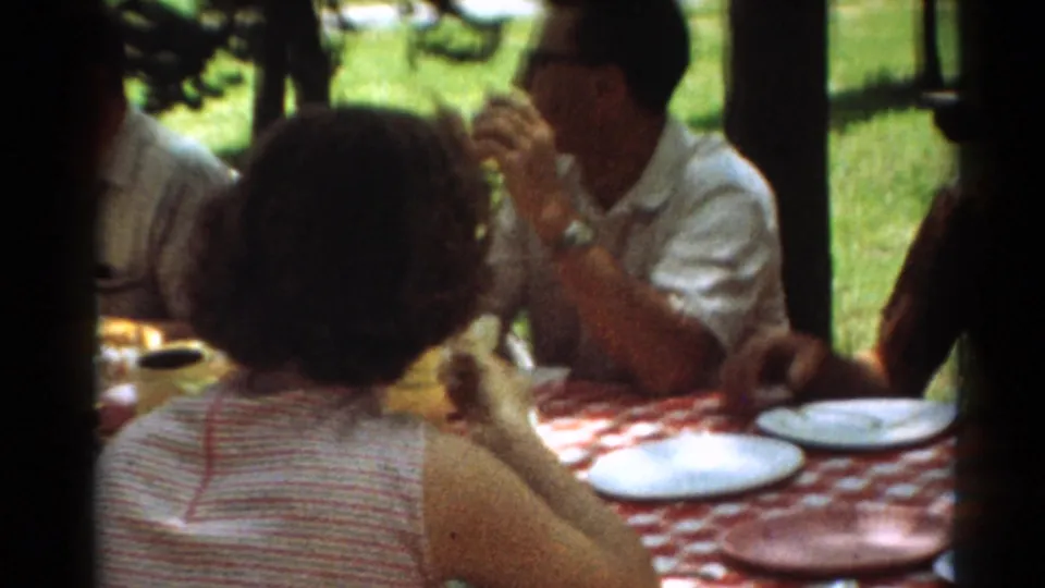 1964 Toledo frame showing a man eating at a shaded picnic table with plates and a red patterned cloth.