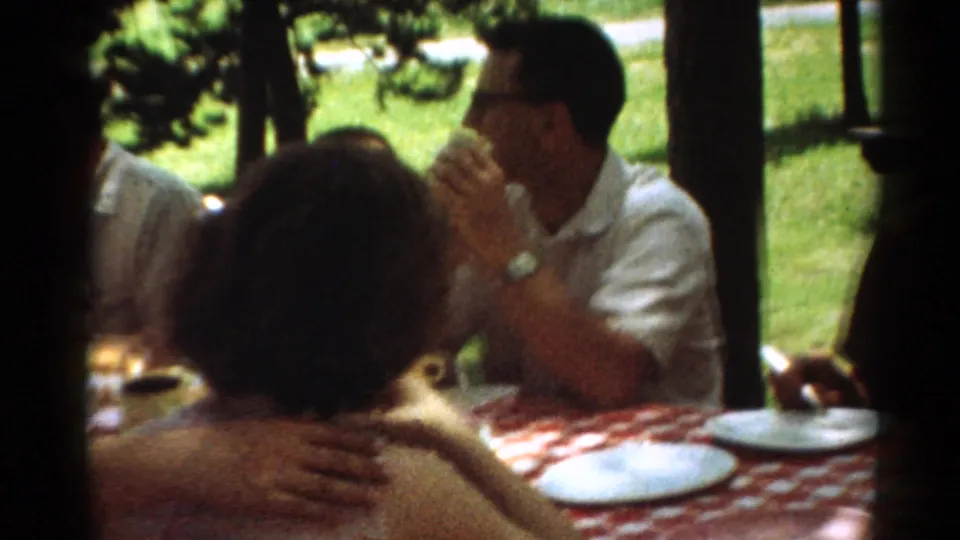 1964 Toledo home-movie frame of adults at a picnic table, including a man in sunglasses beside white plates.