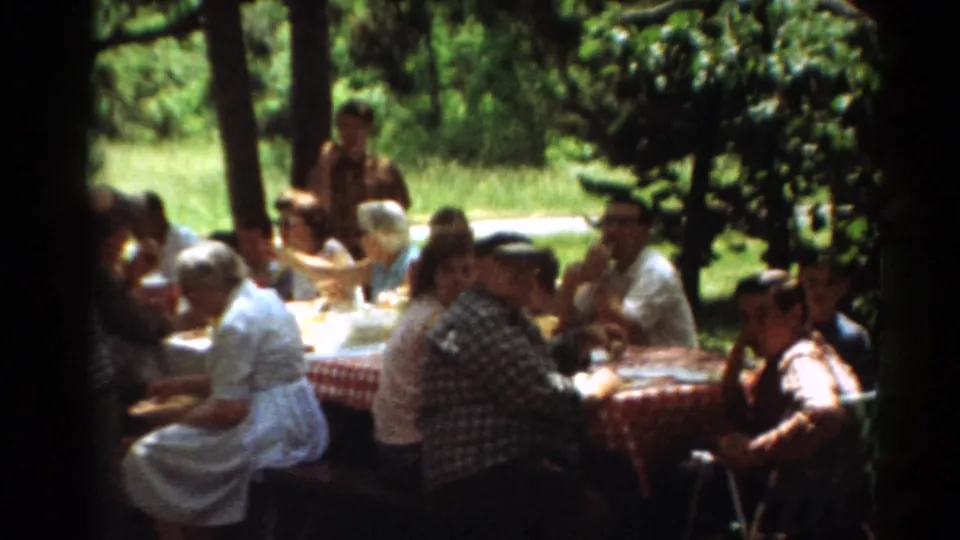 1964 Toledo home-movie frame showing a boy near a checkered picnic table as adults sit and eat in dappled shade.