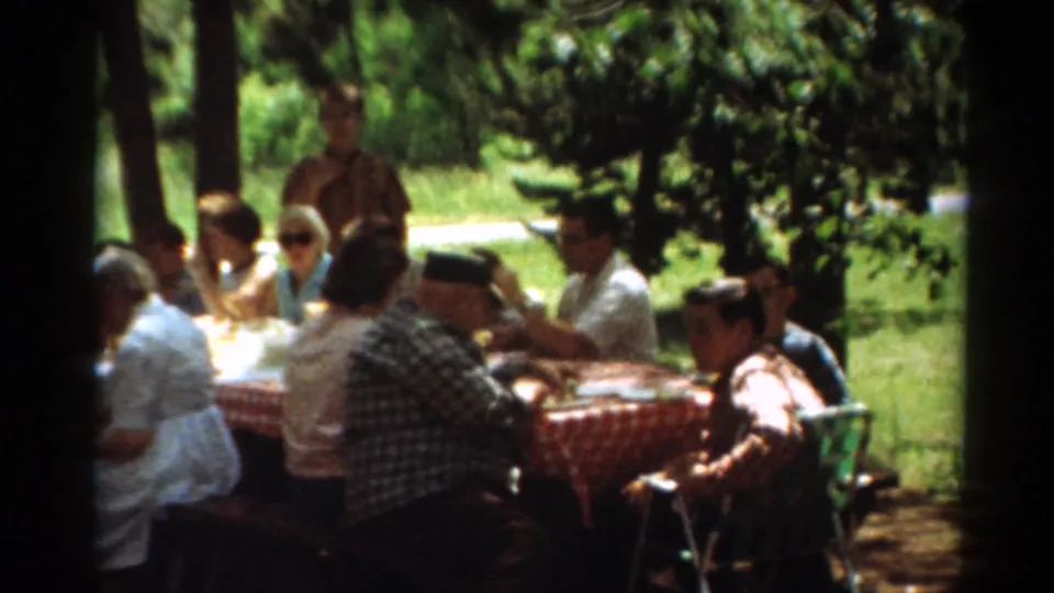 1964 Toledo picnic frame with relatives seated around a red checkered table in a shaded outdoor setting.