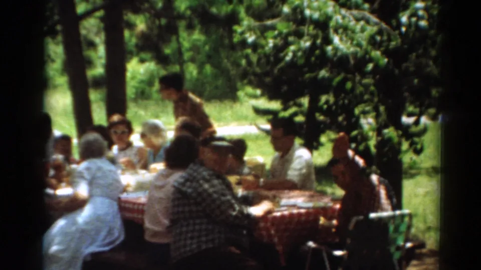 1964 Toledo park picnic frame with seated adults, food, folding chairs, and dappled shade from trees.