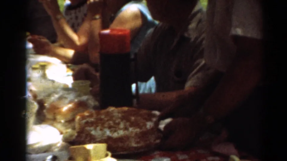 Close 8mm frame of a picnic table with cake, cups, food containers, and adults eating at the 1964 Toledo gathering.