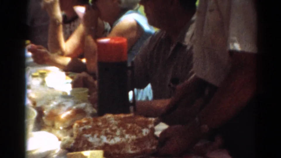 Close frame from the Toledo picnic shows adults seated beside a cake, red thermos, cups, and tableware in strong shade.