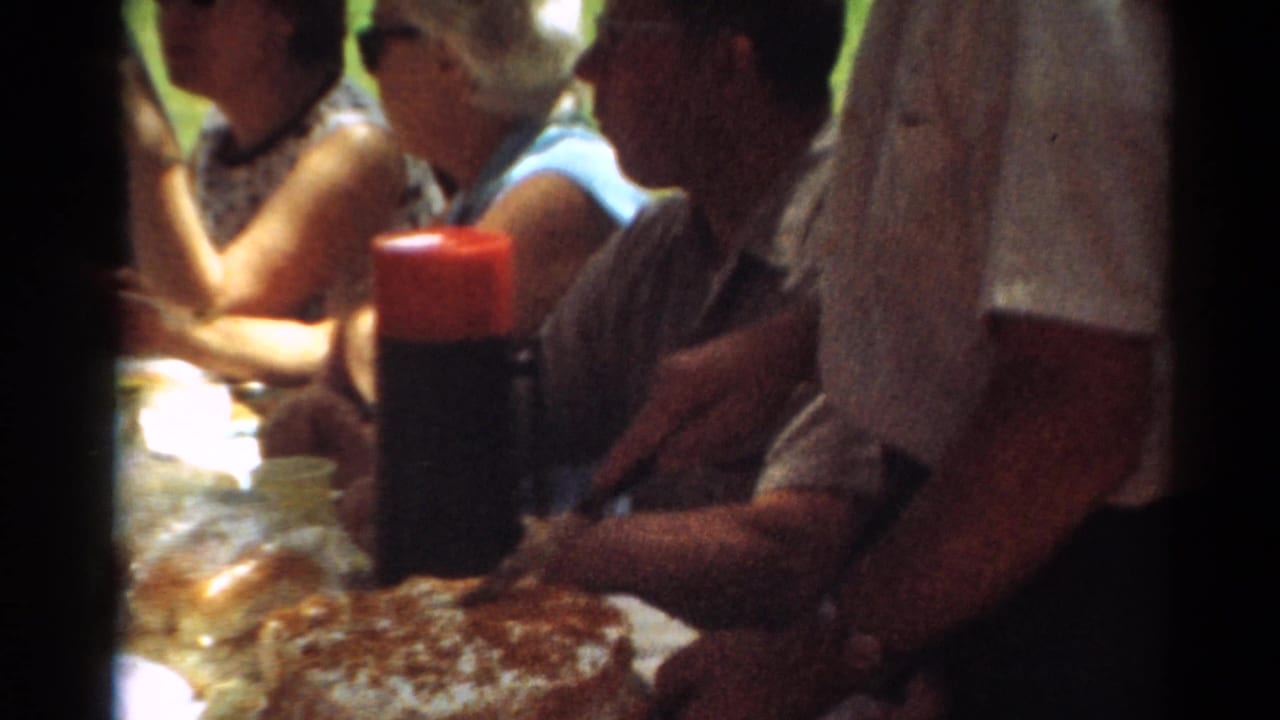 1964 Toledo home-movie frame showing adults seated at a shaded picnic table with food, a red-capped thermos, grass, and trees nearby.