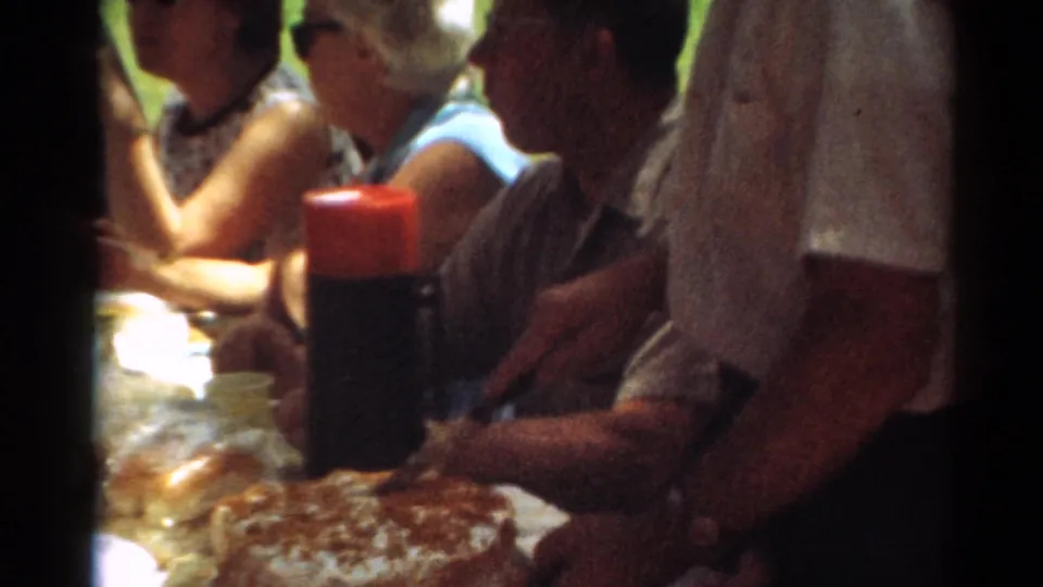 1964 Toledo home-movie frame showing adults seated at a shaded picnic table with food, a red-capped thermos, grass, and trees nearby.