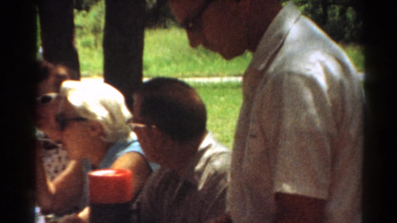 1964 Toledo frame with a woman in sunglasses, a seated man, and a standing man in a light shirt beside a picnic table outdoors.