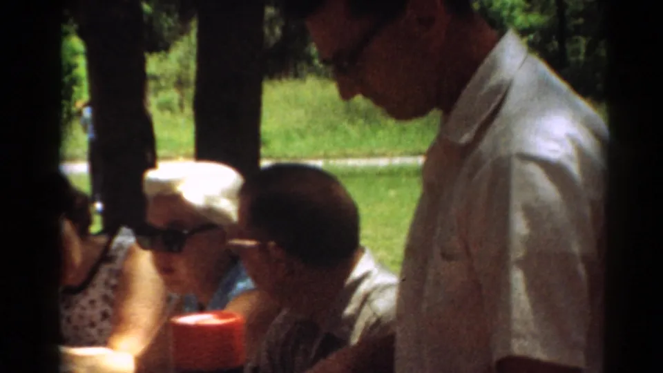 1964 Toledo home-movie frame showing a man standing near seated adults, with a red-capped thermos and outdoor greenery in view.
