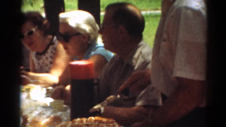 1964 Toledo frame showing adults around a picnic table, a red-capped thermos, dessert on the table, and a standing man handling food.