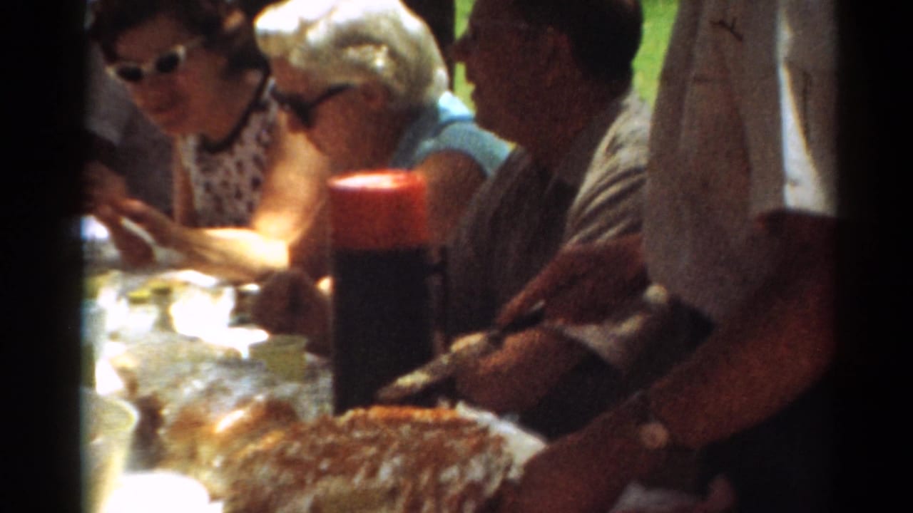 Adults sit at a picnic table with food and a large cake in the foreground.
