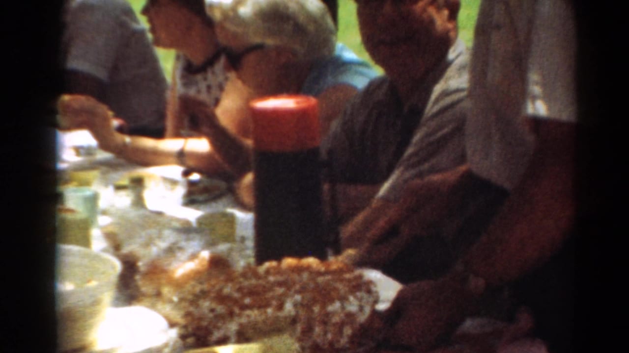 Adults at a 1964 Toledo park picnic sit beside food, cups, and a cake in a warm 8mm frame.