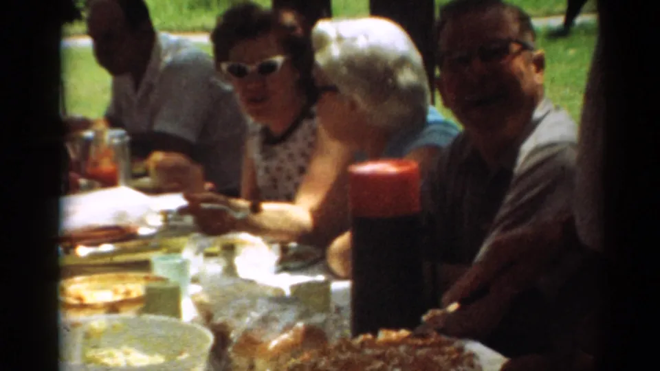 1964 Toledo picnic frame with adults seated at a park table, including a woman in sunglasses and patterned dress.
