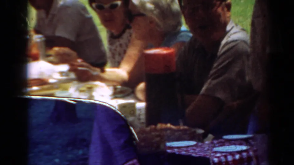 1964 Toledo home-movie frame showing adults at a park picnic table with a checkered cloth and food nearby.