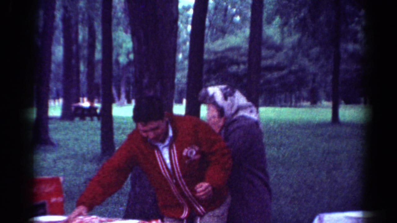 1964 Toledo park picnic frame with a man in a red cardigan and a woman in a headscarf arranging items on a table among trees.
