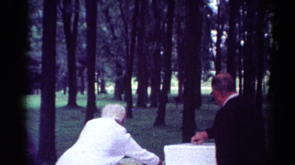 1964 Toledo home-movie frame showing a woman in white and a man in a dark suit at a park picnic table.