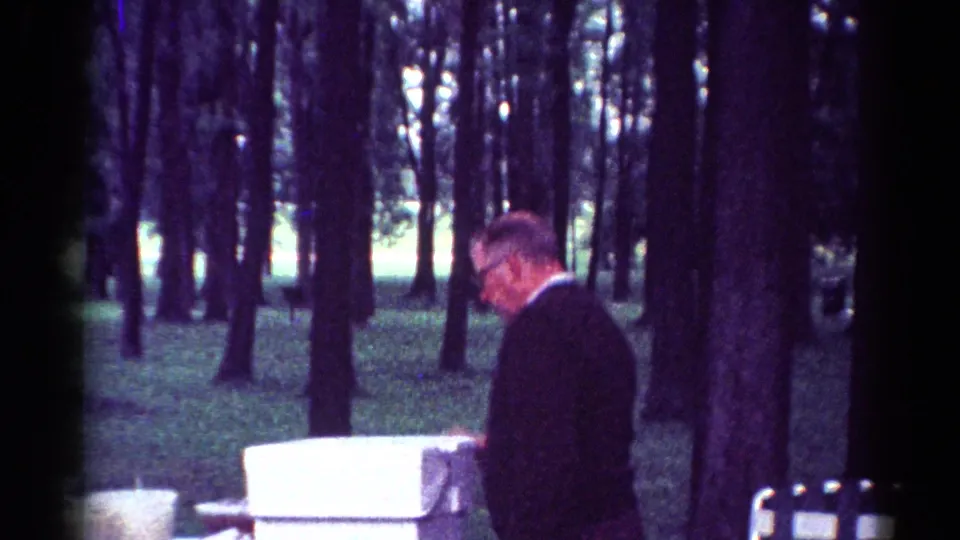 1964 Toledo park picnic frame with a suited man beside white containers and tall trees.