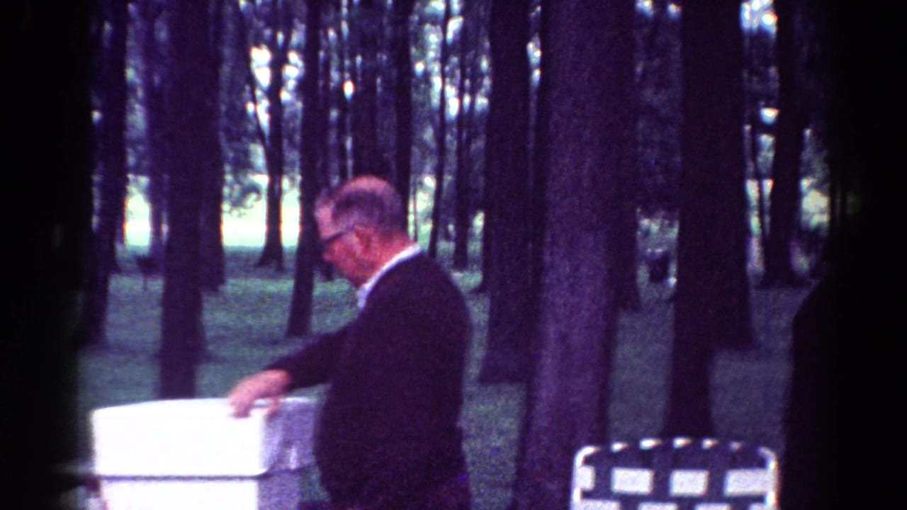 Older man in a dark suit reaches toward a white container at a picnic table in a wooded park.