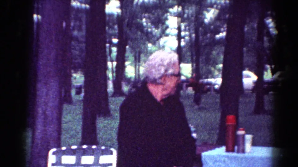 An older woman with white hair and glasses stands at a picnic table in the 1964 Toledo park footage, with cars visible through trees.