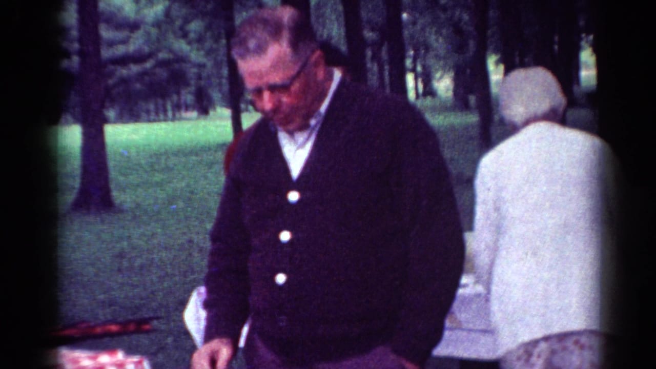 Older man in a dark cardigan stands near a picnic table in a wooded park.